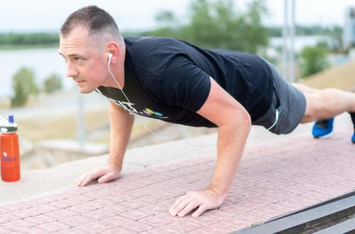 Adult male exercising outdoors performing push-ups with earphones and water bottle.