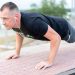Adult male exercising outdoors performing push-ups with earphones and water bottle.